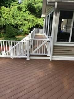 White railing and gate on a deck, leading to a screened porch. Brown deck boards. Green foliage in background.