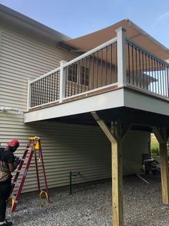 Deck being built with a worker on a ladder, house exterior, gravel ground, and blue sky.