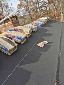 Bags of roofing material arranged in a row on a black surface, near a brick chimney and trees.
