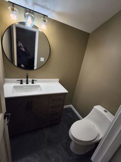 Small bathroom with dark brown vanity, round mirror, white sink, and toilet; beige walls, dark gray floor.