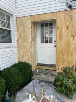 Doorway boarded up with plywood on a white-sided house. Gray steps lead to the door, surrounded by bushes.