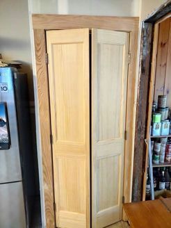 Two wooden, bi-fold doors, closed, in a light-colored wooden frame. Refrigerator and shelves visible.
