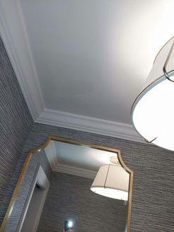 Gold-framed mirror reflects a white lampshade and ceiling. Wallpapered wall.