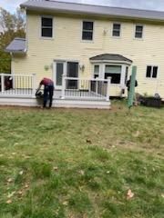 Person trimming grass near a yellow house with a deck. Green grass, blue sky.