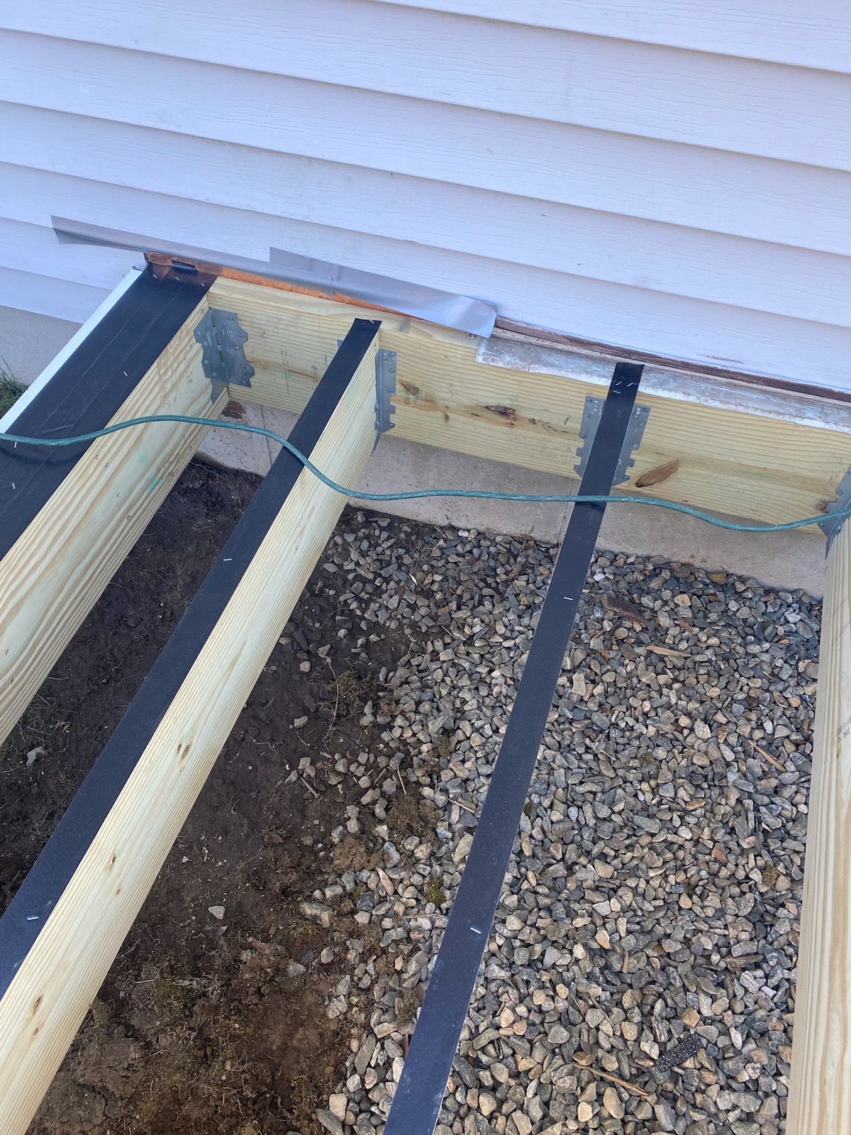 Wooden deck frame under construction with gravel base and house siding.