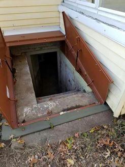 Brown cellar doors open to a concrete stairway leading into a dark basement entrance on the side of a house.