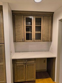 Built-in desk with wooden cabinets; glass-fronted upper cabinets, white backsplash, light countertop.