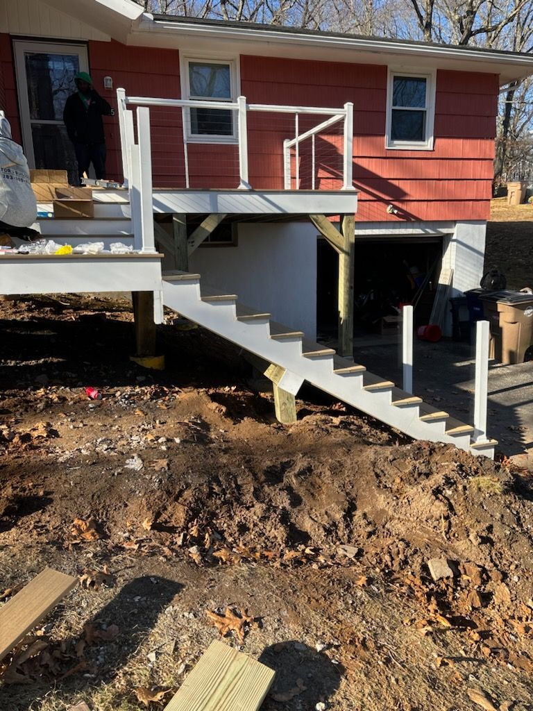 A partially constructed deck with stairs attached to a red house, built above a dirt yard.