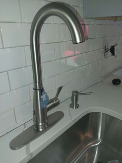 Stainless steel kitchen faucet with sprayer and soap dispenser, against white tiled backsplash.