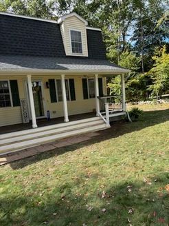 Yellow house with a black roof and porch; green lawn and trees in background.