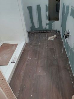 Bathroom under construction, with wood-look tile flooring partially installed. White walls and shower base visible.