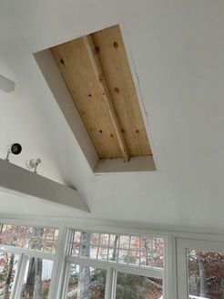 Rectangular opening in white ceiling reveals wood framing. Sunroom interior with windows, lights.