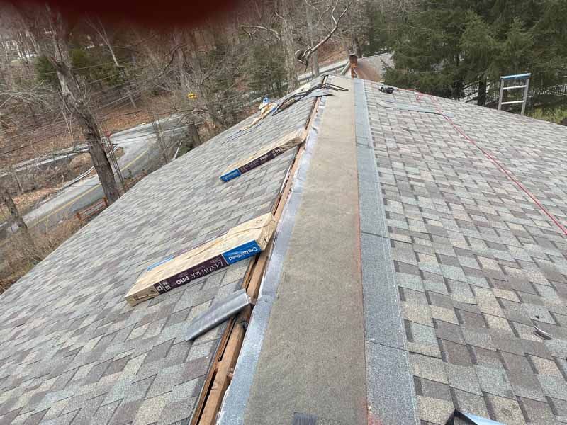 Rooftop with asphalt shingles being worked on; bundles of shingles, safety line, and ladder visible.