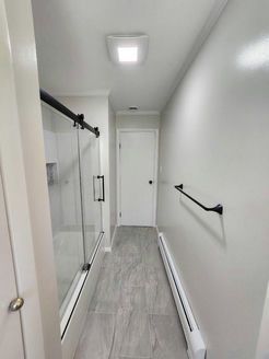 Narrow bathroom with gray tile floor, glass shower, white door, and black towel rack.