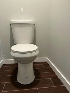 White toilet in a small corner bathroom with dark brown tile floor and gray walls.