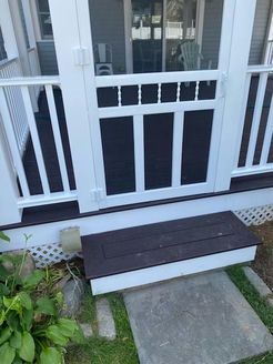 White screen door and porch railing with dark brown steps, grass and concrete path.