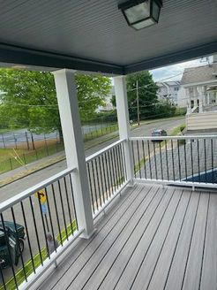 A grey and white porch overlooking a street, with black railing and green trees in the background.