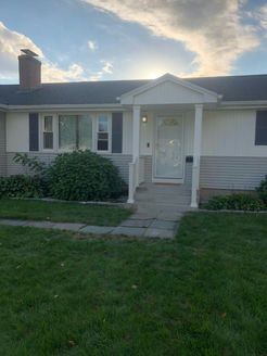 Single-story home with a white door, porch, and a brick chimney. Green lawn and a cloudy sky.
