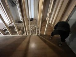 Person crouches near wooden framing; debris fills the space behind.