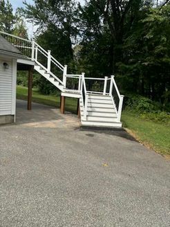 White wooden stairs and railing lead up from asphalt driveway to a deck attached to a white building.
