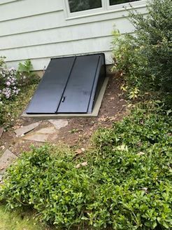 Black basement door on a sloped concrete base, set into a white house exterior with surrounding greenery.