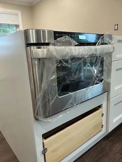 Stainless steel built-in oven, wrapped in plastic, installed in white kitchen cabinetry. A drawer is below it.