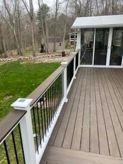 Wooden deck with white railing, black spindles, and a glass-enclosed sunroom. Green grass and trees in background.