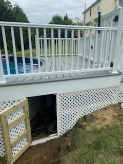 White deck with lattice skirting, access door open to storage, overlooking a pool.