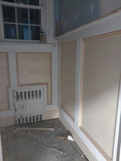 Interior view of a room under renovation. Newly installed wall panels with molding, window, and radiator visible.
