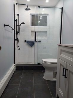 Modern bathroom with black-framed shower, white tile, black fixtures, and a toilet. Light blue walls and dark floor tiles.