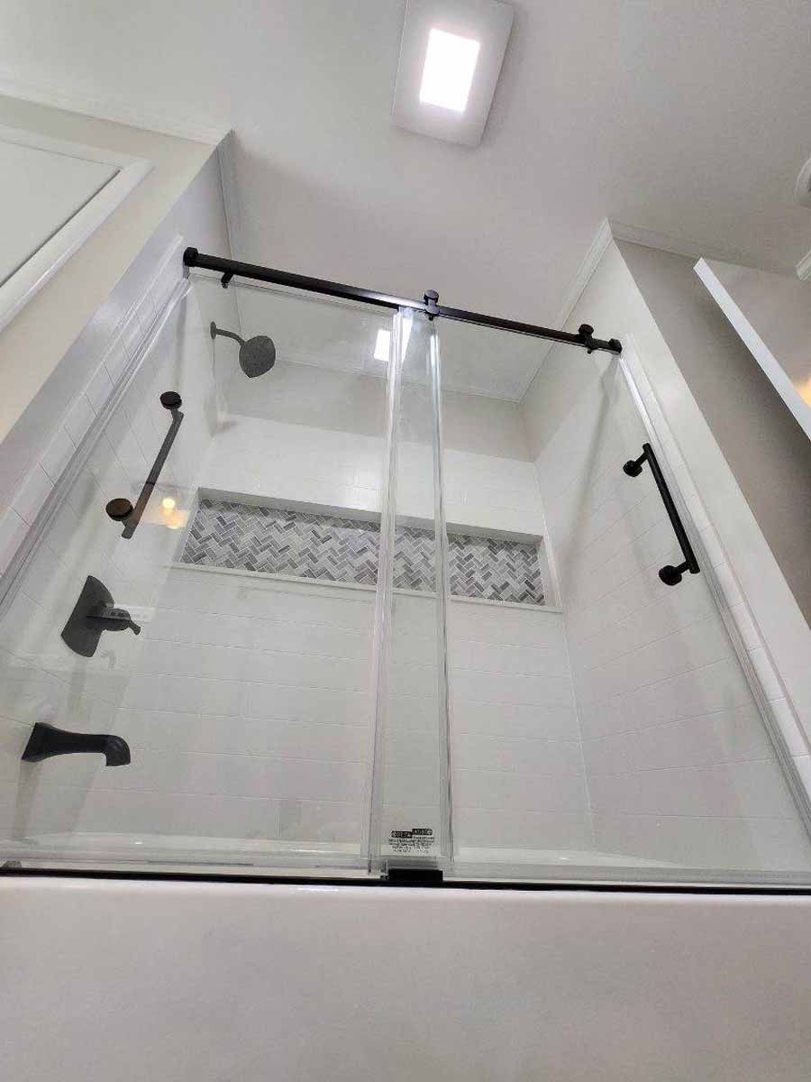 A modern shower with clear glass sliding doors and black hardware. White tile walls with a mosaic accent.