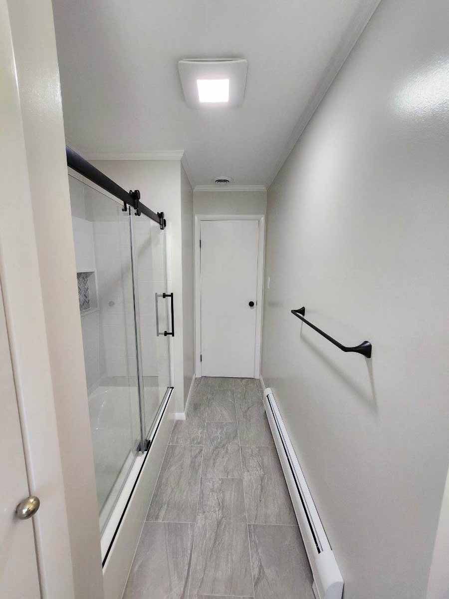 Narrow bathroom with gray tile floor, glass shower, white walls, and a closed white door.