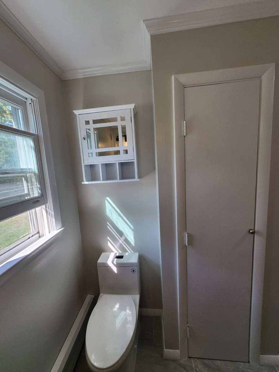 Small bathroom with toilet, white wall cabinet, door, and window. Light gray walls and white trim.