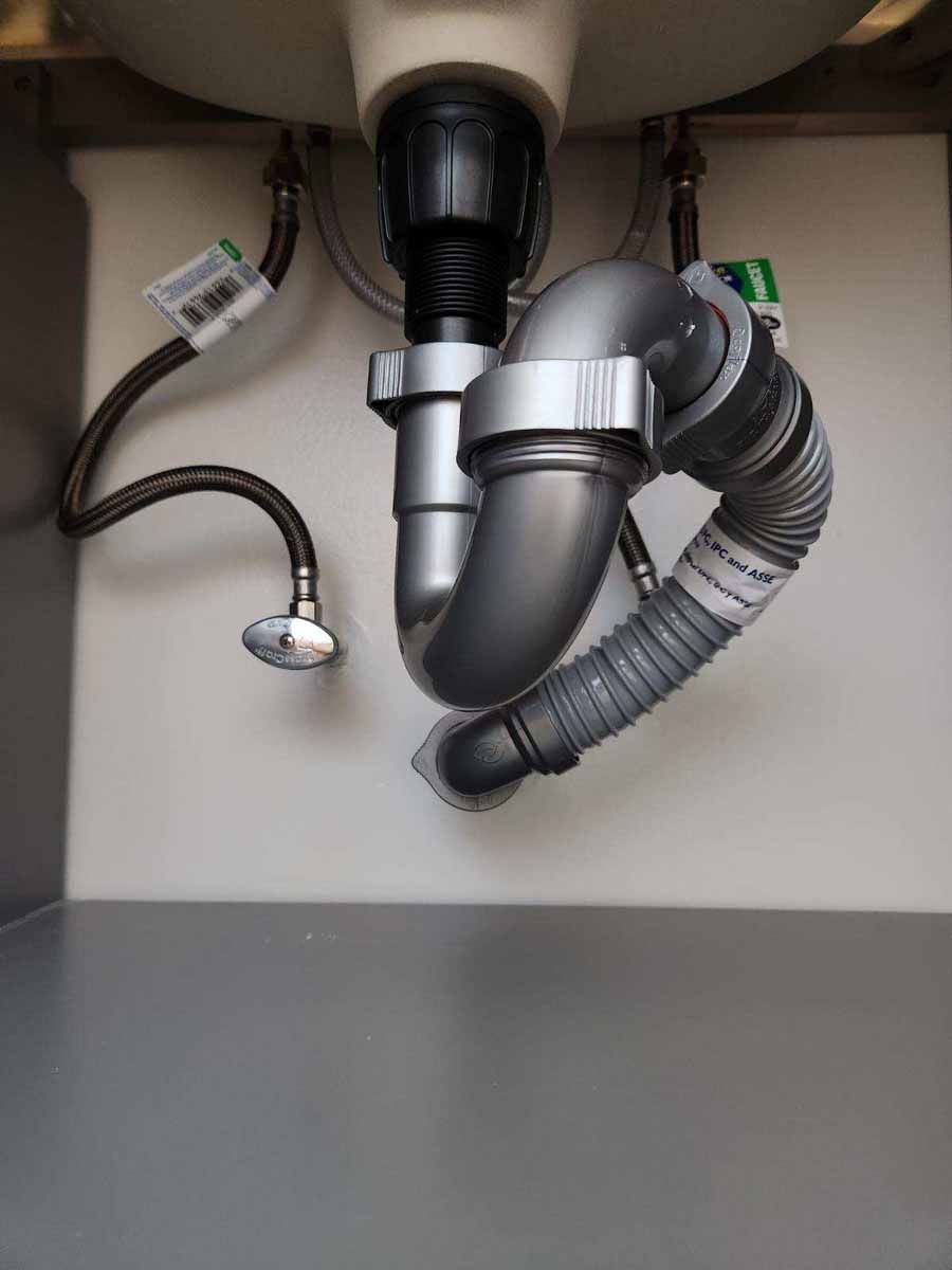 Under-sink plumbing with pipes and a flexible drain tube, white cabinet interior, gray floor.