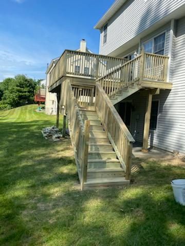 Wooden deck and stairs attached to a two-story house, set in a grassy yard under a blue sky.