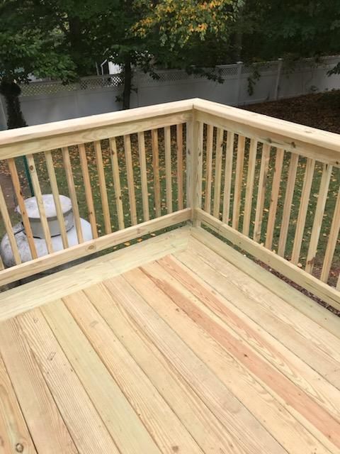 Wooden deck with railing, constructed outdoors; green trees and white fence in background.