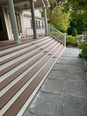 Brown and cream deck stairs leading to a white porch with a stone walkway alongside, surrounded by greenery.
