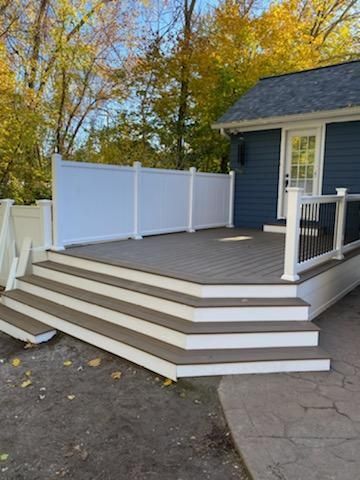 Gray deck with white railings and steps against a blue house. Autumn trees in the background.