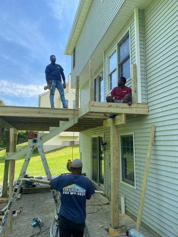 Construction workers building a wooden deck on a house. One worker stands on the deck, another sits, and a third works below.