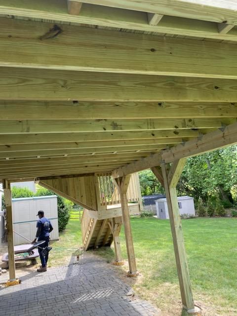 A person works under a new wooden deck on a brick patio. Green yard and sheds are in the background.