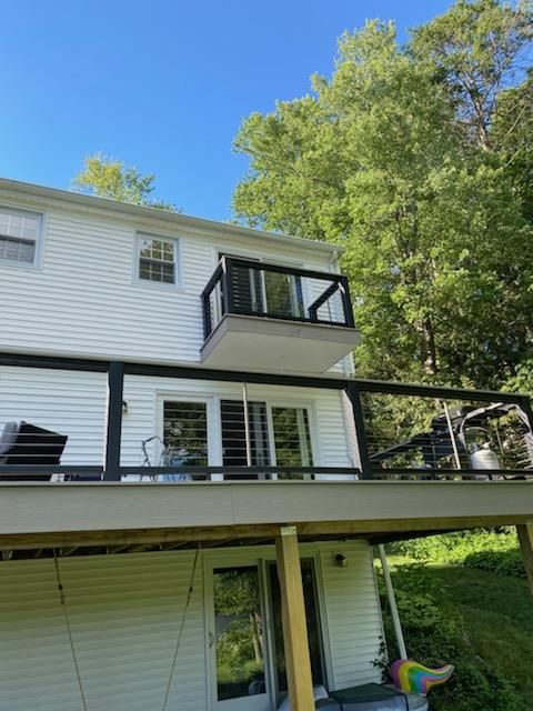 White house with multiple decks, black railings, and glass panels. Lush green trees in the background.