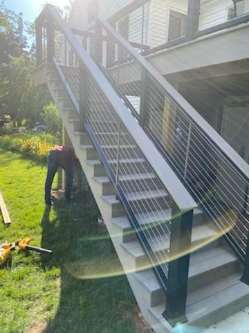 Outdoor staircase with gray steps and metal cable railing, constructed beside a house on a grassy lawn.