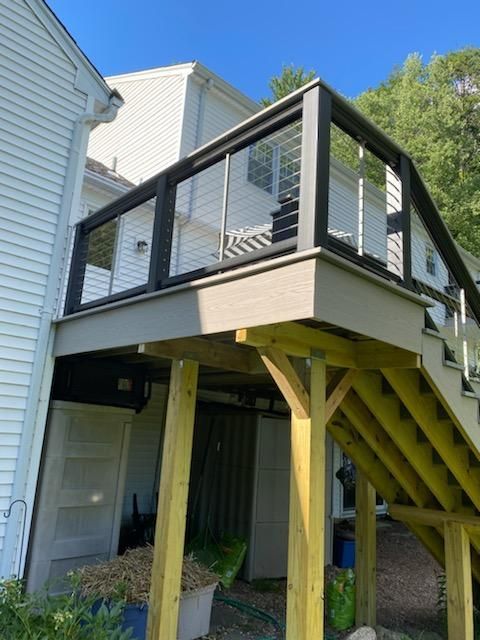 Deck with black railing, cable infill, and stairs, attached to a white house with a blue sky background.