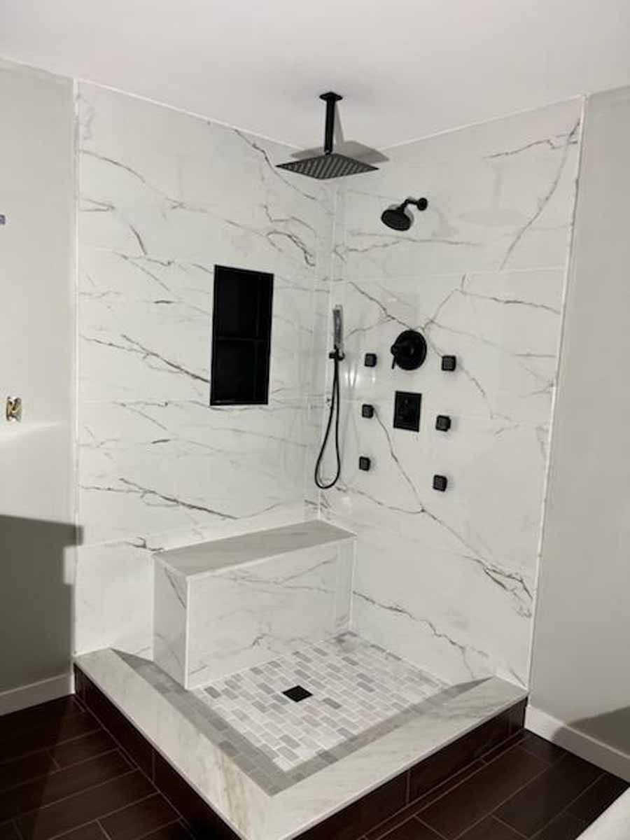 Modern marble-tiled shower with a bench, multiple showerheads, and dark fixtures in a corner setting.