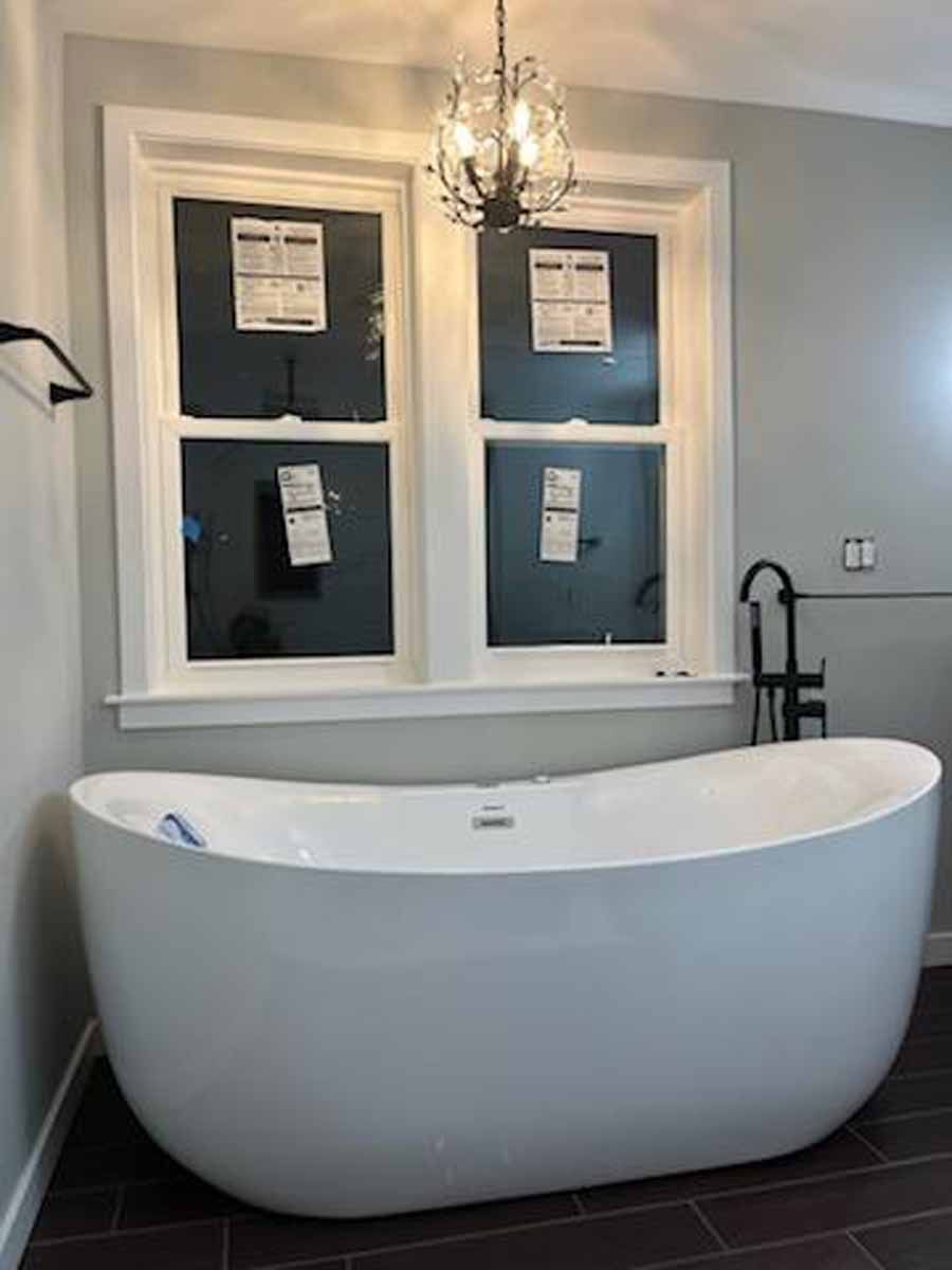 Freestanding white bathtub under a window in a bathroom, dark floor, gray walls, chandelier.