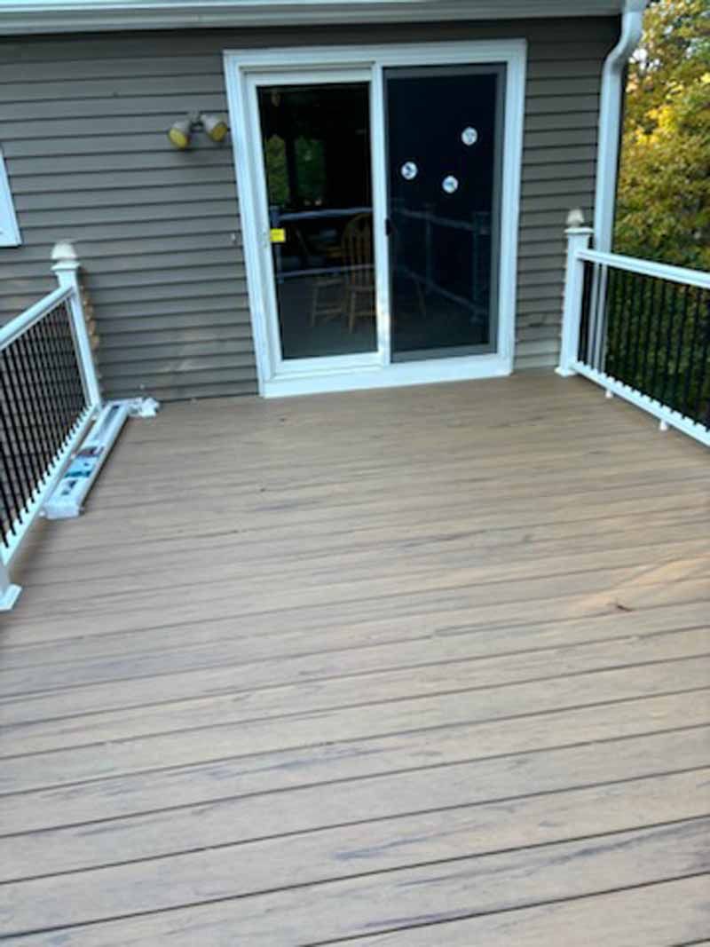 A deck with light brown planks, a sliding glass door, and white railing, leading to a house with gray siding.