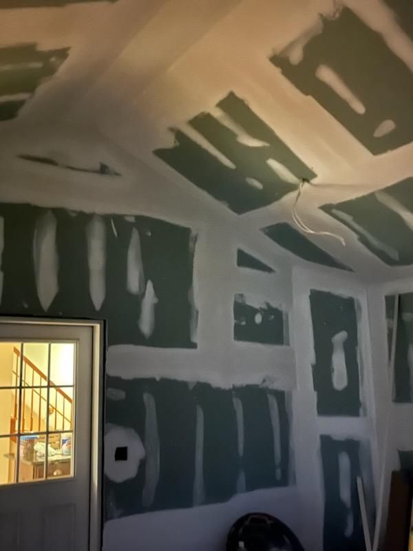 Interior of a room with unfinished drywall installation, showing green walls and white ceiling. A door and wiring are visible.