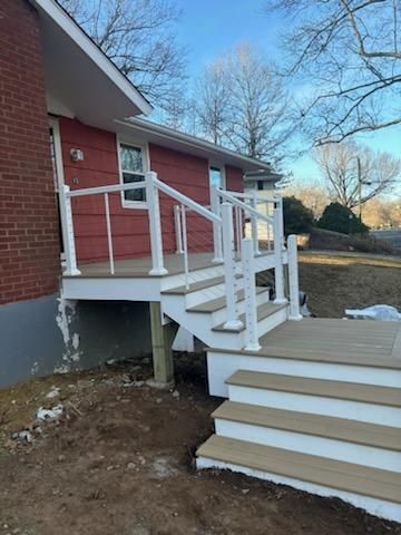 Red house with white railing deck and stairs, built in yard with bare trees.