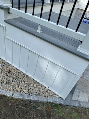 White-paneled deck skirting with a gray deck top and black railing, over gravel and stone pavers.
