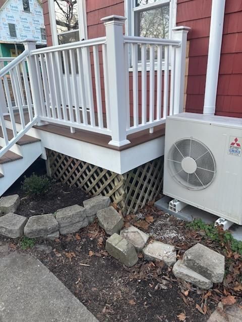 A red house with a white railing and steps. A heat pump is next to it, and the ground has stone edging.